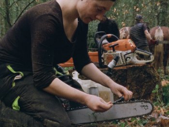 Copyright: Filmgarten Person schärft im Wald die Kette einer Motorsäge, im Hintergrund führt eine weitere Person ein Zugpferd mit Rückewagen.