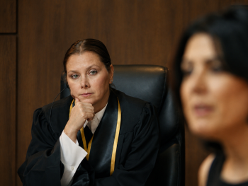 Copyright: The Center for Human Rights in Iran
Eine Richterin in Robe sitzt nachdenklich im Gerichtssaal, während im Vordergrund unscharf eine weitere Person zu sehen ist.