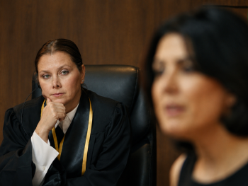 Copyright: The Center for Human Rights in Iran Eine Richterin in Robe sitzt nachdenklich im Gerichtssaal, während im Vordergrund unscharf eine weitere Person zu sehen ist.