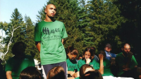 Eine stehende Person in grünem „Seeds of Peace International Camp“-T-Shirt spricht zu einer Gruppe sitzender Personen in gleicher Kleidung im Freien vor Bäumen.