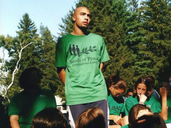 Copyright: Temps noir Eine stehende Person in grünem „Seeds of Peace International Camp“-T-Shirt spricht zu einer Gruppe sitzender Personen in gleicher Kleidung im Freien vor Bäumen.