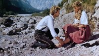 Ein junger Mann in Tracht kniet an einem Fluss und hält den Fuß einer jungen Frau, die auf einem Stein sitzt, vor einem Bergpanorama.