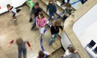 Kinder laufen und spielen aus der Vogelperspektive über eine Treppe mit einem Bodenband, auf dem Begriffe wie „Lesen“, „Mailen“ und „Schauen“ stehen.