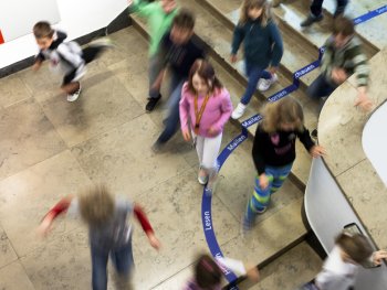 Kinder laufen und spielen aus der Vogelperspektive über eine Treppe mit einem Bodenband, auf dem Begriffe wie „Lesen“, „Mailen“ und „Schauen“ stehen.