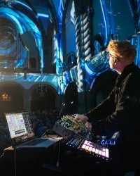 Der Soundkünstler Paul Biessmann steht mit Laptop und Mischpult in einer großen Halle, die mit blauem Licht beleuchtet wird.