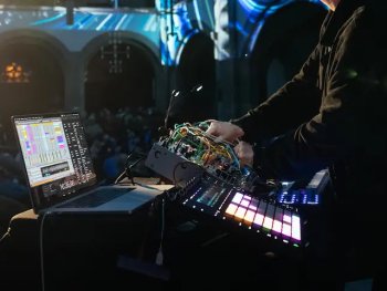 Der Soundkünstler Paul Biessmann steht mit Laptop und Mischpult in einer großen Halle, die mit blauem Licht beleuchtet wird.