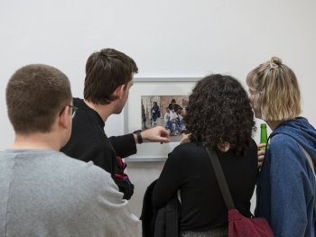Vier Personen betrachten von hinten ein gerahmtes Foto einer Personengruppe an einer weißen Wand, eine Person zeigt dabei auf das Bild.