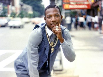 Foto: Jamel Shabazz, NYC 1981
Ein Mann in hellblauem Nadelstreifenanzug mit goldenen Ketten und Ringen posiert lächelnd auf einem Gehweg in einer Stadtstraße.