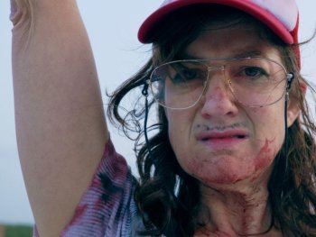 Copyright: filmgalerie451
Person mit roter Kappe und Brille, blutverschmiertem Gesicht und erhobenem Arm blickt ernst in die Kamera.