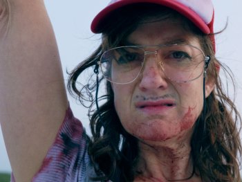 Copyright: filmgalerie451 Person mit roter Kappe und Brille, blutverschmiertem Gesicht und erhobenem Arm blickt ernst in die Kamera.