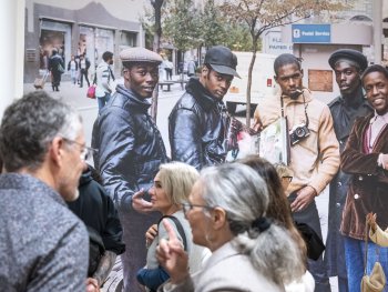 Ausstellungsbesucherinnen und -besucher stehen im Gespräch vor einem großformatigen Wandfoto, das fünf Männer mit Kameras und Fotoalbum auf einer Straße zeigt.