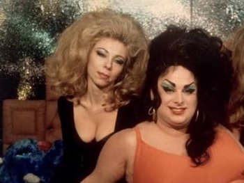 Copyright: Filmverleih
Drei Personen mit toupierten Frisuren und auffälligem Make-up posieren in glamourösen Outfits vor einem glitzernden Hintergrund.