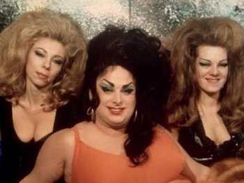 Copyright: Filmverleih Drei Personen mit toupierten Frisuren und auffälligem Make-up posieren in glamourösen Outfits vor einem glitzernden Hintergrund.