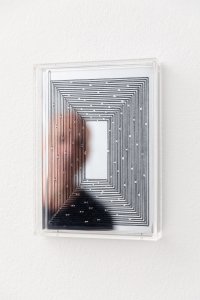 Gerahmtes Kunstwerk mit konzentrischem Linienmuster auf spiegelnder Oberfläche, in dem sich eine unscharf zu sehendePerson spiegelt.