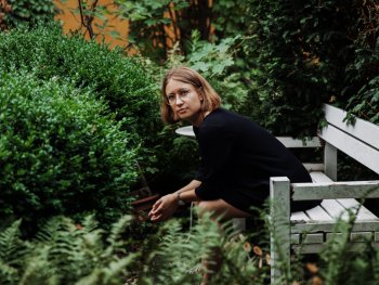 Foto: Dovile Sermokas
Rebecca Trescher sitzt auf einer weißen Holzbank inmitten dichter grüner Pflanzen und blickt in die Kamera.