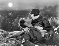 Schwarz-Weiß-Filmszene, in der ein Mann im Gras liegt und eine Frau mit Hut ihm nahe an das Gesicht gebeugt ist, im Hintergrund Schilf und Mond.
