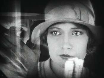 Copyright: Friedrich-Wilhelm-Murnau-Stiftung Schwarz-weiße Filmaufnahme einer Person mit Glockenhut in Nahaufnahme, die ein weißes Tuch an den Mund hält und mit ernstem Blick in die Kamera schaut.