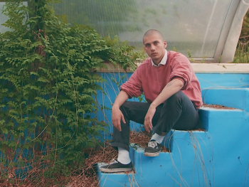 Eine Person mit rasiertem Kopf und rosafarbenem Pullover sitzt auf blauen Stufen neben wuchernden Pflanzen in einem verlassenen Gewächshaus.