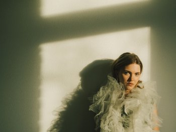 Foto: Tim Cavadini
Eine Person in einem Kleid mit Tüll-Rüschen lehnt an einer Wand im Licht- und Schattenspiel eines Fensters.