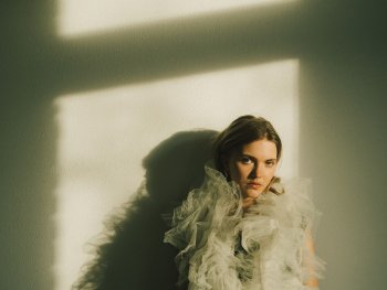 Eine Person in einem Kleid mit Tüll-Rüschen lehnt an einer Wand im Licht- und Schattenspiel eines Fensters.