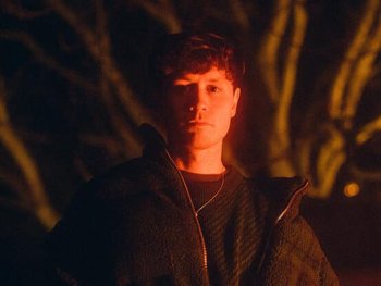 Eine Person steht nachts vor kahlen Bäumen und wird von warmem, orangefarbenem Licht angestrahlt.