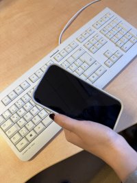 Eine Hand hält ein Smartphone mit schwarzem Display über einer weißen Computertastatur auf einem Schreibtisch.