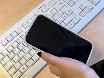 Copyright: Künstlerhaus Eine Hand hält ein Smartphone mit schwarzem Display über einer weißen Computertastatur auf einem Schreibtisch.