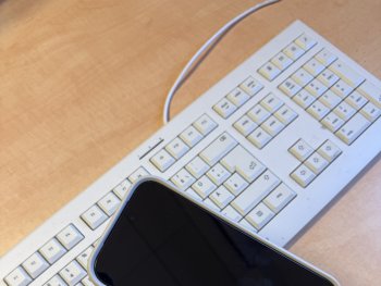 Foto: Künstlerhaus
Eine Hand hält ein Smartphone mit schwarzem Display über einer weißen Computertastatur auf einem Schreibtisch.
