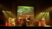 Musiker spielen auf einer beleuchteten Bühne mit Schlagzeug, Kontrabass, Saxofon und Vibrafon, während im Hintergrund eine zeichnerische Landschaftsprojektion zu sehen ist.