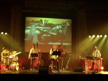 Musiker spielen auf einer beleuchteten Bühne mit Schlagzeug, Kontrabass, Saxofon und Vibrafon, während im Hintergrund eine zeichnerische Landschaftsprojektion zu sehen ist.