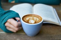Eine Hand hält eine Tasse Kaffee mit Latte-Art vor einem aufgeschlagenen Buch auf einem Holztisch.