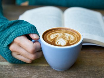 Eine Hand hält eine Tasse Kaffee mit Latte-Art vor einem aufgeschlagenen Buch auf einem Holztisch.