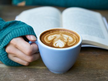 Eine Hand hält eine Tasse Kaffee mit Latte-Art vor einem aufgeschlagenen Buch auf einem Holztisch.