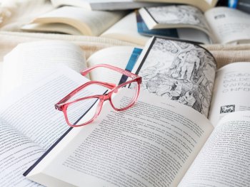 Eine rosa Brille liegt auf einem aufgeschlagenen Buch inmitten mehrerer geöffneter Bücher mit bedruckten Seiten.