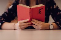 Person hält ein aufgeschlagenes rotes Buch vor sich und liest an einem Tisch sitzend.