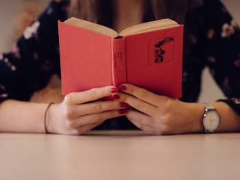 Person hält ein aufgeschlagenes rotes Buch vor sich und liest an einem Tisch sitzend.
