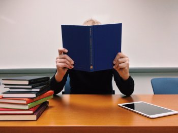 Eine Person hält ein aufgeschlagenes blaues Buch vor dem Gesicht, während an einem Tisch daneben ein Bücherstapel und ein Tablet liegen.