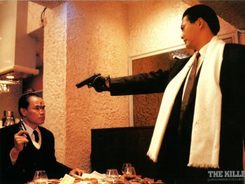 Copyright: 2026 Plaion Pictures; REM
Zwei Männer in Anzügen richten im Filmstill zu „The Killer“ Pistolen aufeinander, während ein verletzter Mann am Tisch sitzt.