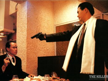 Copyright: 2026 Plaion Pictures; REM Zwei Männer in Anzügen richten im Filmstill zu „The Killer“ Pistolen aufeinander, während ein verletzter Mann am Tisch sitzt.