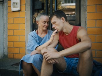 Copyright: Grandfilm Zwei Personen sitzen vor einem Hauseingang mit gelber Ziegelfassade und führen ein nachdenkliches Gespräch.