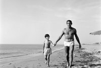Schwarz-Weiß-Foto eines Mannes und eines Jungen in Badehosen, die händchenhaltend am Strand entlang spazieren.