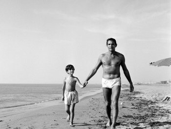 Copyright: Filmverleih Schwarz-Weiß-Foto eines Mannes und eines Jungen in Badehosen, die händchenhaltend am Strand entlang spazieren.