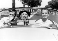 Drei Männer sitzen in einem offenen Cabriolet, der Mann in der Mitte trägt einen Hut und raucht Pfeife, im Hintergrund eine von Bäumen gesäumte Landstraße.