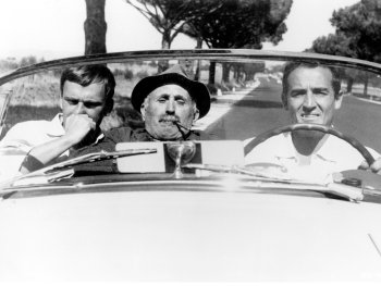 Copyright: Filmverleih Drei Männer sitzen in einem offenen Cabriolet, der Mann in der Mitte trägt einen Hut und raucht Pfeife, im Hintergrund eine von Bäumen gesäumte Landstraße.