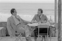 Zwei Männer in Anzügen sitzen an einem Tisch mit Getränken auf einer Terrasse am Meer und unterhalten sich.