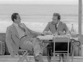 Copyright: Filmverleih Zwei Männer in Anzügen sitzen an einem Tisch mit Getränken auf einer Terrasse am Meer und unterhalten sich.