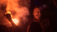 Ein Mann mit geöffnetem Mund steht bei Nacht neben einer brennenden Fackel mit Funkenflug und Rauch.