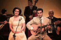 Eine Sängerin und ein Gitarrist im Stil der 1950er Jahre sitzen vor Mikrofonen auf einer Bühne, umgeben von einer Band mit Kontrabass und Akkordeon.