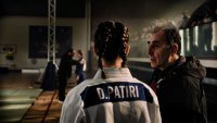 Eine Judoka mit geflochtenen Haaren im weißen Judo-Anzug spricht mit einem älteren Mann am Rand einer Wettkampfmatte.
