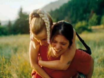 Copyright: Salzgeber und Co. Medien Eine lachende Person trägt ein Kind huckepack auf einer grünen Wiese vor bewaldeten Bergen.